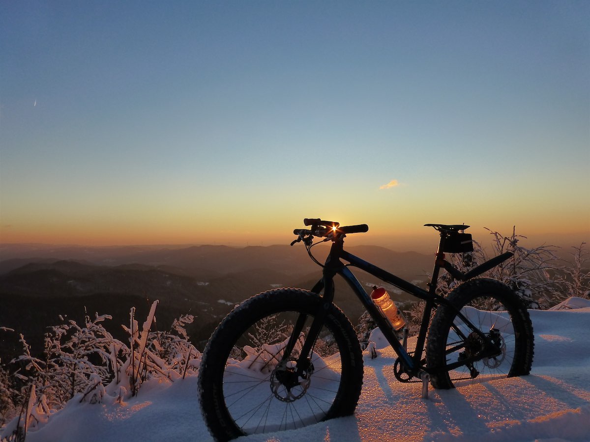 bikerumor pic of the day a fat bike sits in th snow on the edge of a mountain looking out over the darkened land below as the sun sets.