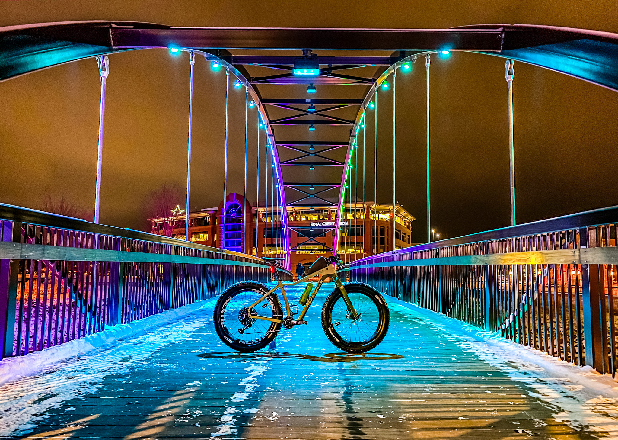 Bikerumor pic of the day Eau Claire, Wisconsin, a bicycle is placed in the center of a walking bridge at night, the lights are turquoise and hot purple and seem to glow on the snow covered bridge.