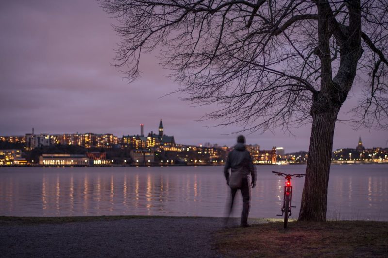 bikerumor pic of the day stockholm sweden a cyclist stands with their bike next to a tree along a river at night with the lights of Södermalm and old town on the other side of the river making the sky light up purple with the clouds