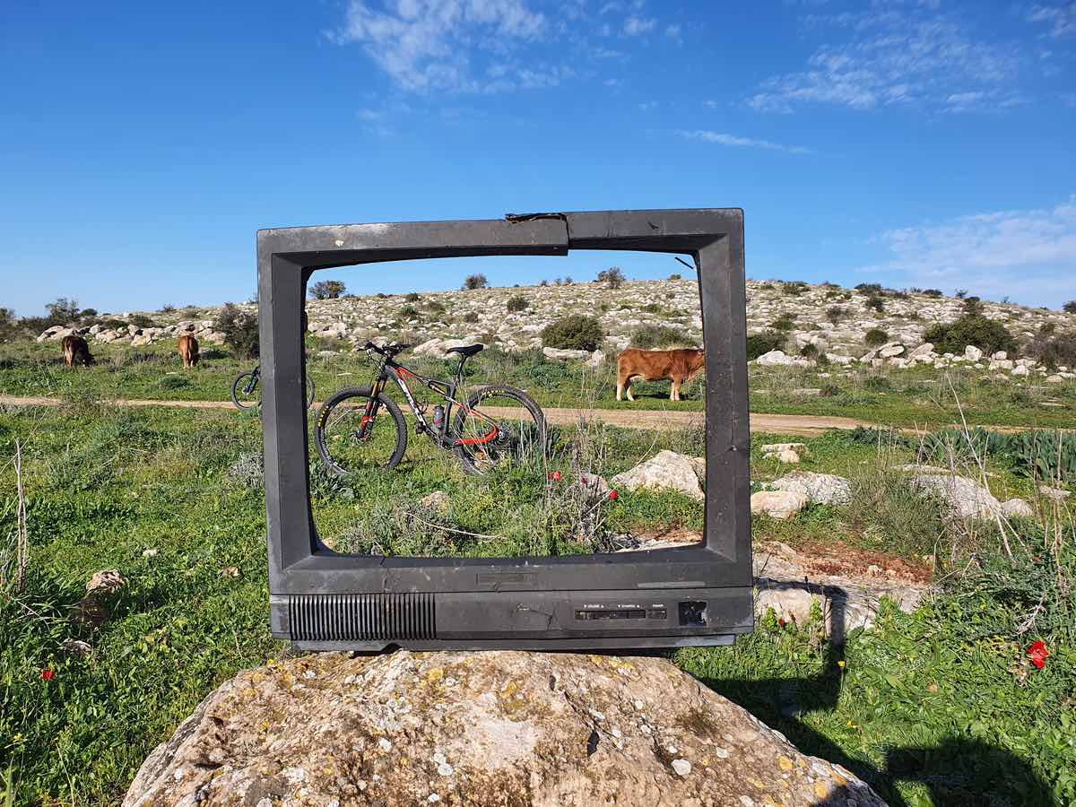 bikerumor pic of the day a photo of a hollowed out tv screen sitting on a rock in a grassy field a bicycle is seen through the tv fram with cows and rocky outcropping in the distance.