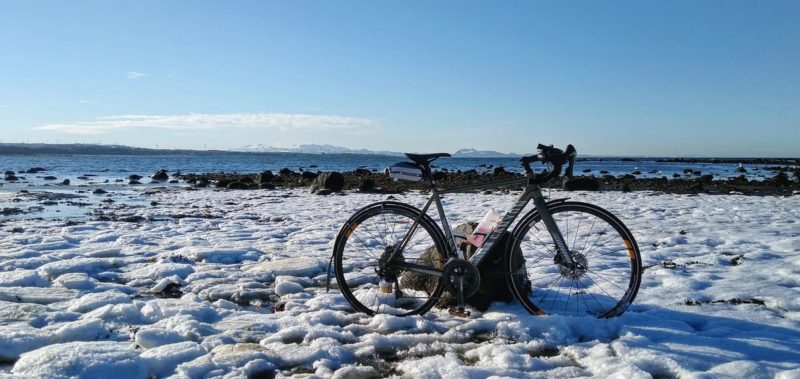 bikerumor pic of the day Longniddry Bents, Scotland, a bicycle is leaning against a small boulder in a light snow with a body of water and land beyond