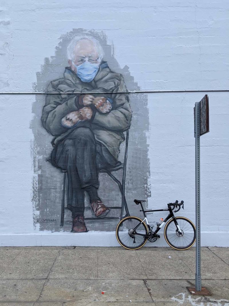 bikerumor pic of the day a photo of a bicycle leaning against a wall with a giant mural of Bernie Sanders and his mittens.