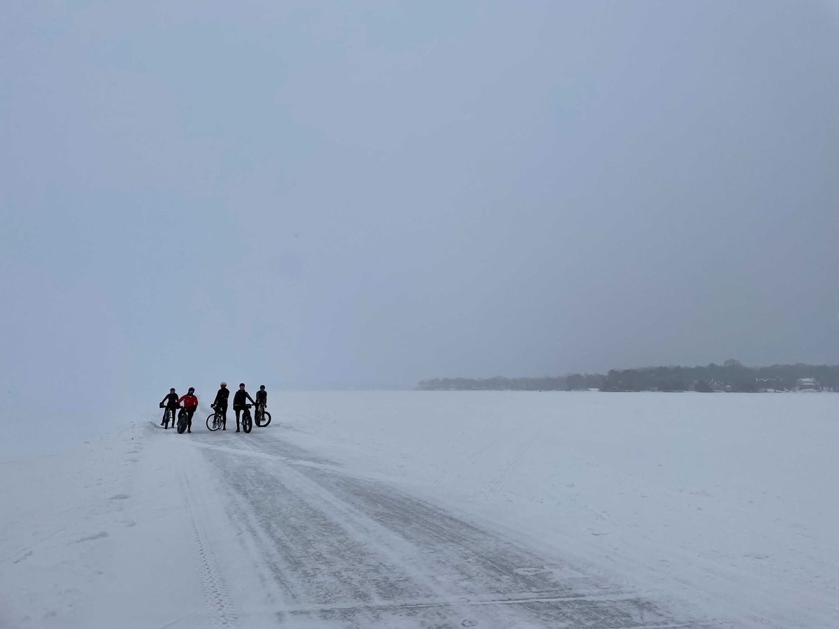 bikerumor pic of the day bone saw cycling collective group of cyclists out on a frozen lake minnetonka, mn, with snow surrounding and a small batch of trees in the distance. The sky is covered in dense grey clouds.