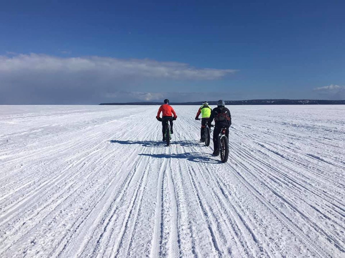 bikerumor pic of the day three cyclists riding across a frozen lake huron to Mackinac Island, there is nothing else around and the sky is blue and sun is shining on the riders.