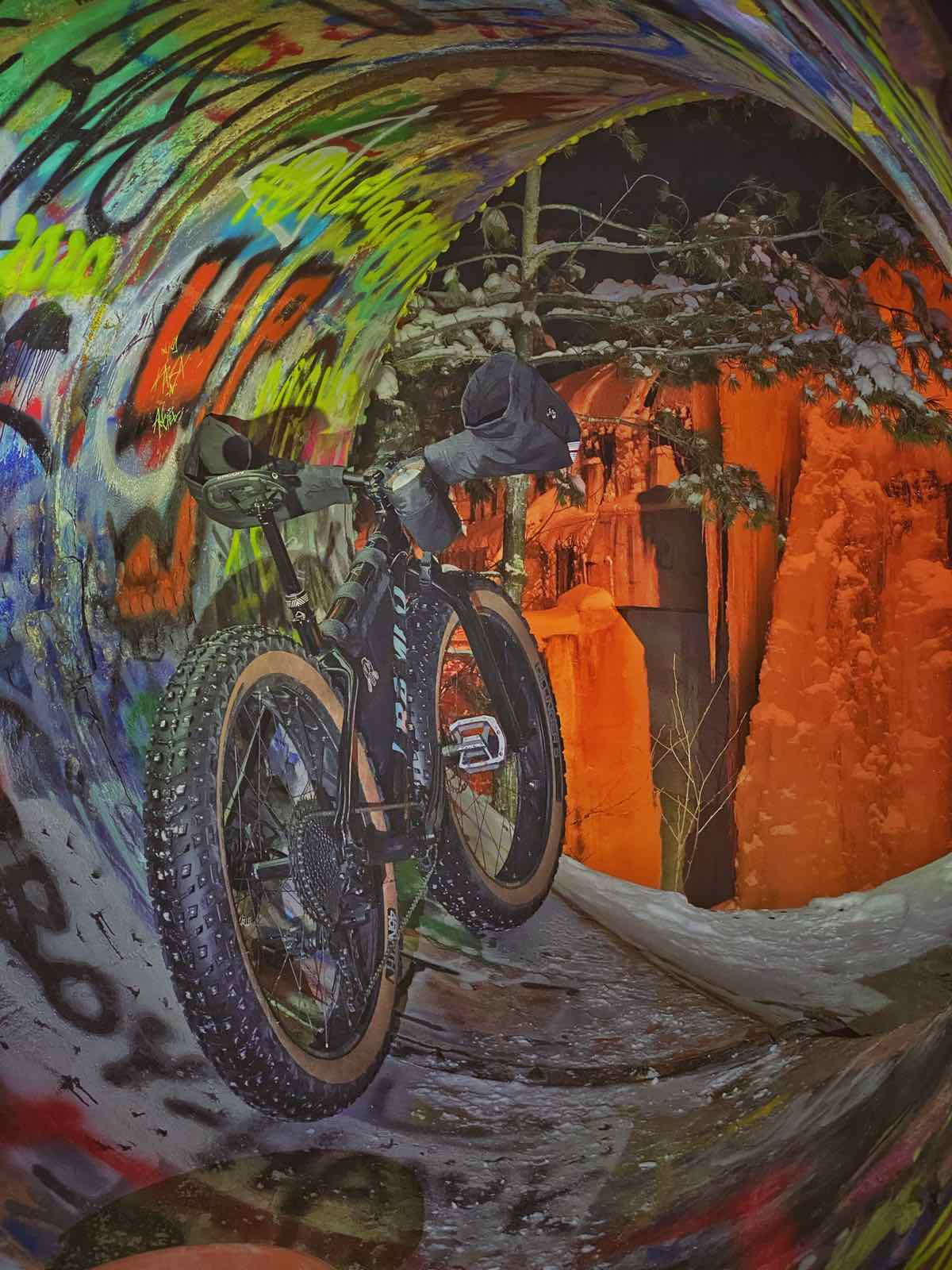 bikerumor pic of the day a fat bike is inside a drainage pipe with colorful graffiti inside and what looks like ice beyond the tunnel with an orange light on it.