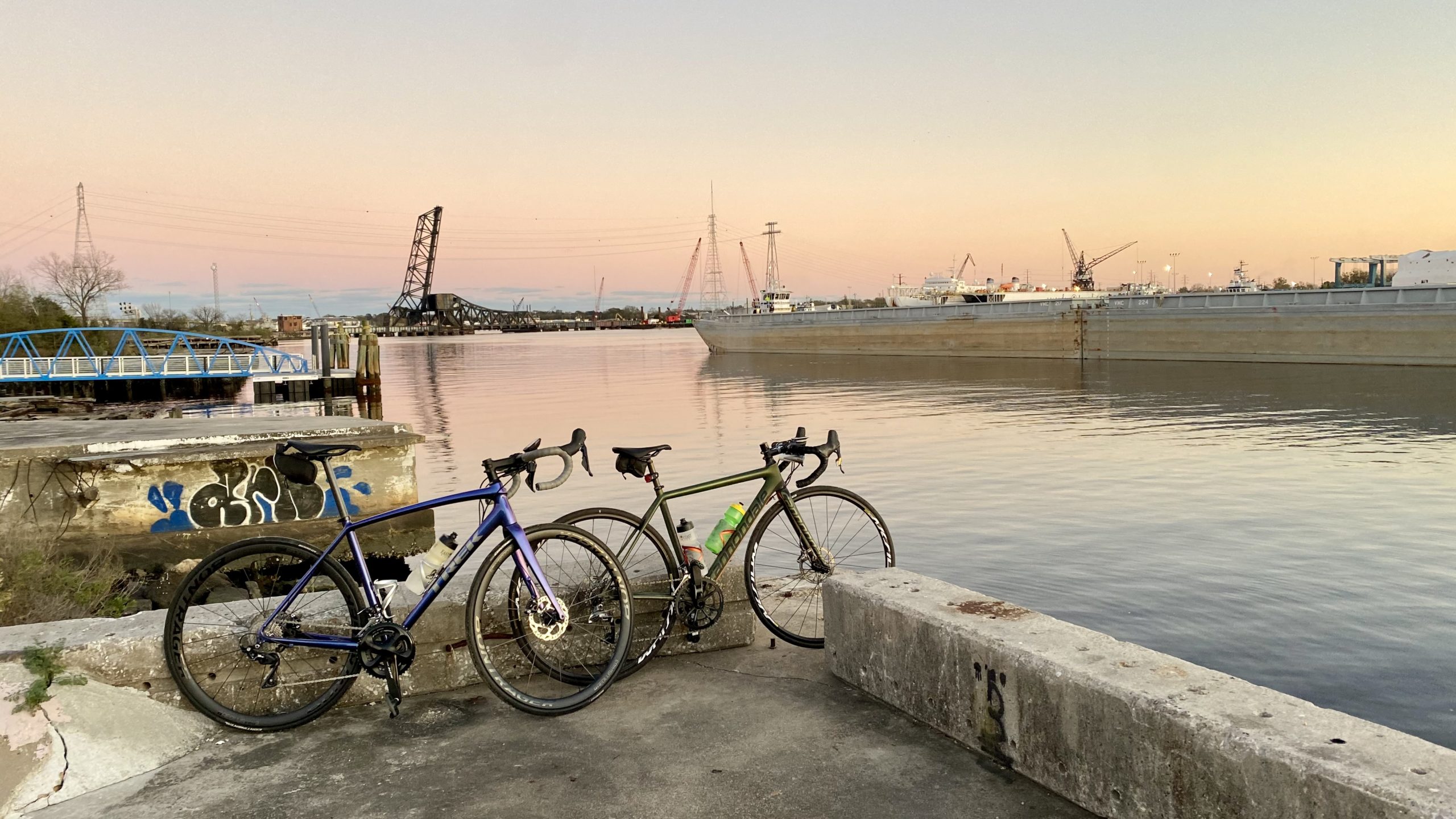 bikerumor pic of the day two bicycles lean against a concrete barrier along the elizabeth river in norfolk virginia as the sun has set and the pink sky is reflecting off the river.