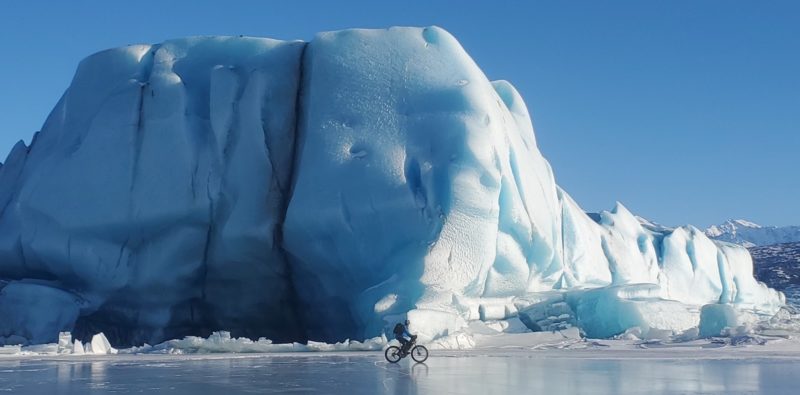 a photo of a mountain biker riding on a frozen lake with a giant glacier behind them. the mountain biker looks very small compared to the glacier.