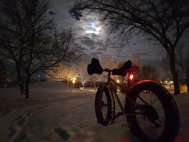 bikerumor pic of the day surly mainlander is in the snow on a snowy trail in the dark pointed towards lights in Lacombe Lake Park in St. Albert AB Canada the wolf moon peeks out from the clouds