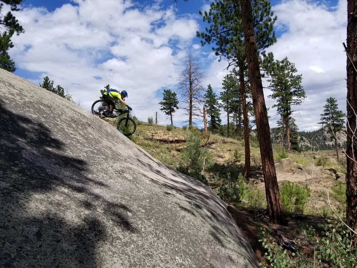 bikerumor pic of the day buffalo creek trails in colorado a mountain biker leans far back on their bike as they ride down a smooth steep rock there are pine trees in the background and the blue sky is dotted with clouds.