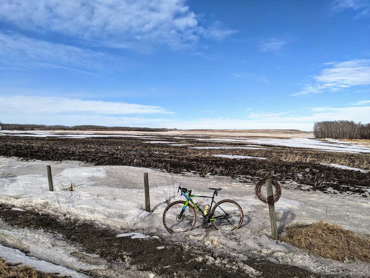 bikerumor pic of the day Sturgeon County, Alberta, bike leaning against a barbed wire fence with snow covering the side of the road and muddy field in the background the sky is blue with some whispy clouts.