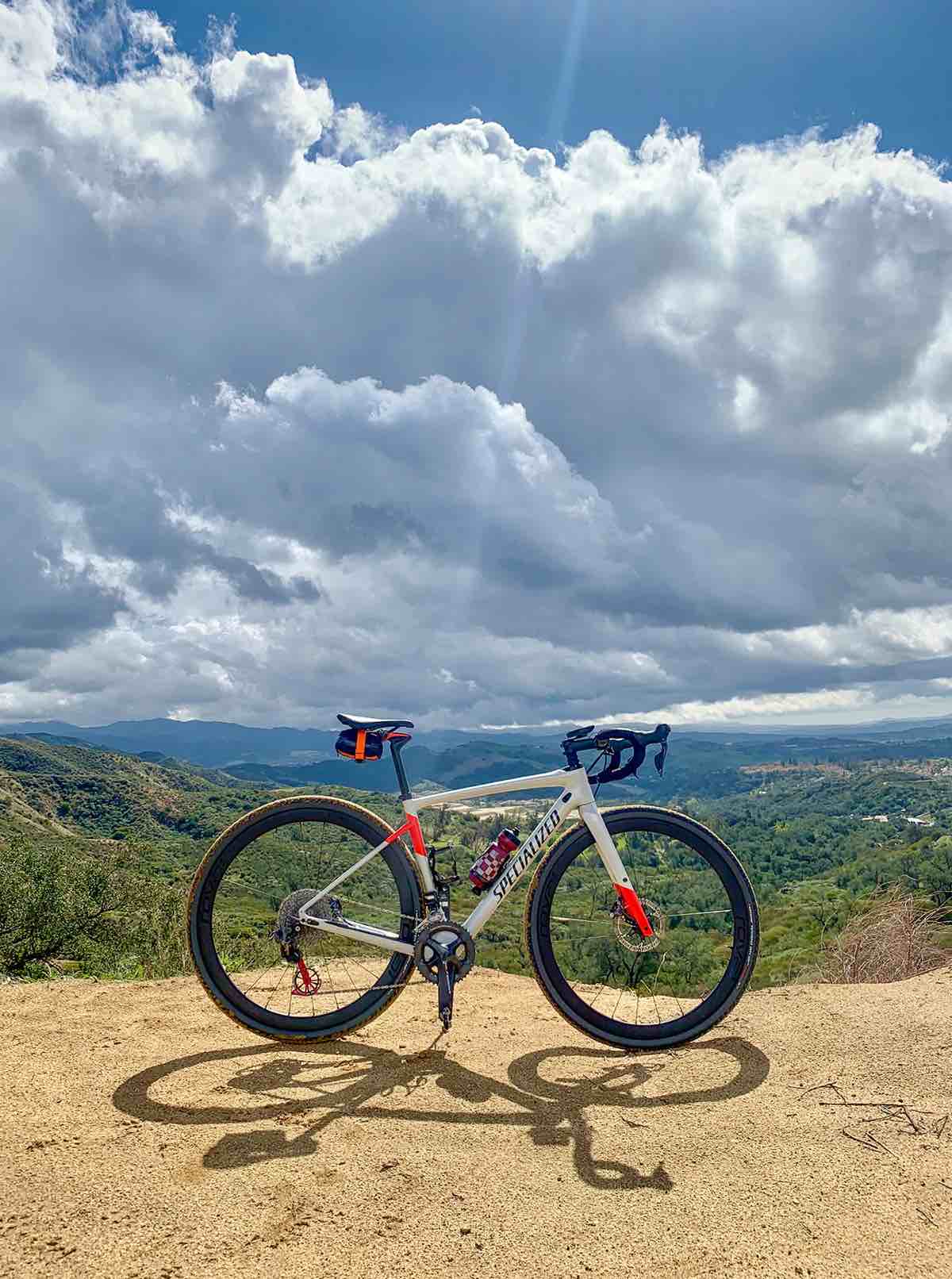 bikerumor pic of the day Trabuco Canyon, CA - Santiago Truck Trail entrance, a bicycle is along a dirt mountain ridge the a cloudy sky in the background and green hills beyond, the sky has opened up above the bicycle and it is lit up with bright sun