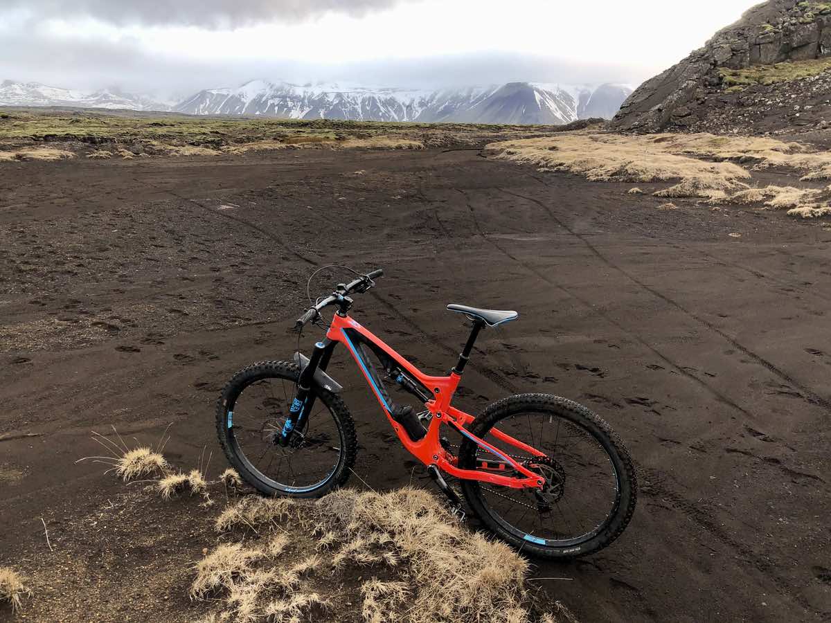 a red mountain bike is in a field of lava rock sand with tall snow capped mountains in the distance in iceland