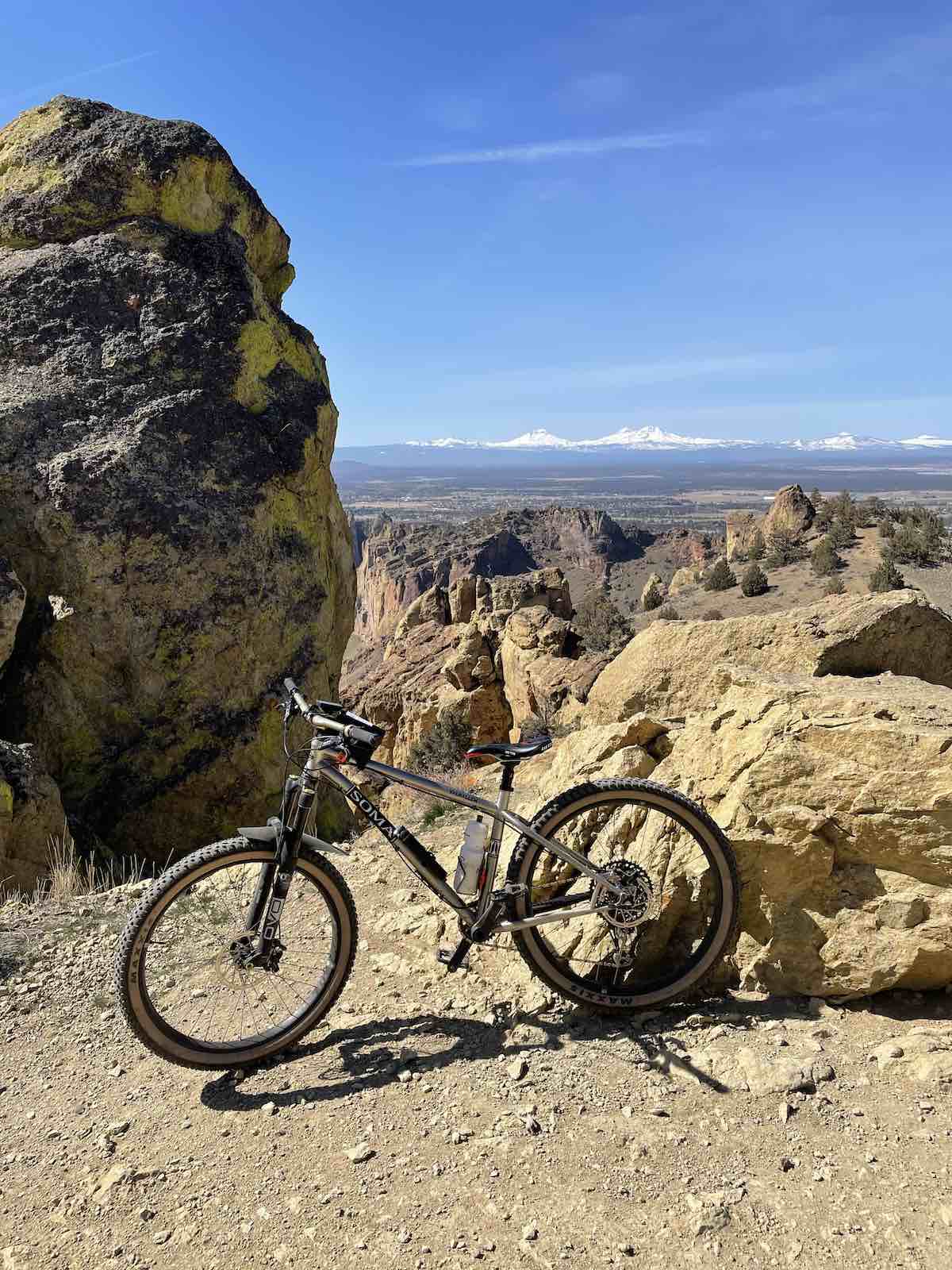 bikerumor pic of the day a mountain bike is posed near a large rock with another larger rock formation to the left of the photo snow capped mountains can be seen in the distance with clear blue skies.