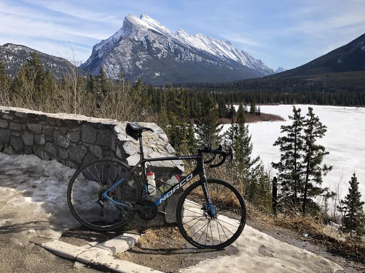 bikerumor pic of the day a bicycle leans against a low stone wall with a lake surrounded by pine trees and a snow capped mountain in the distance.
