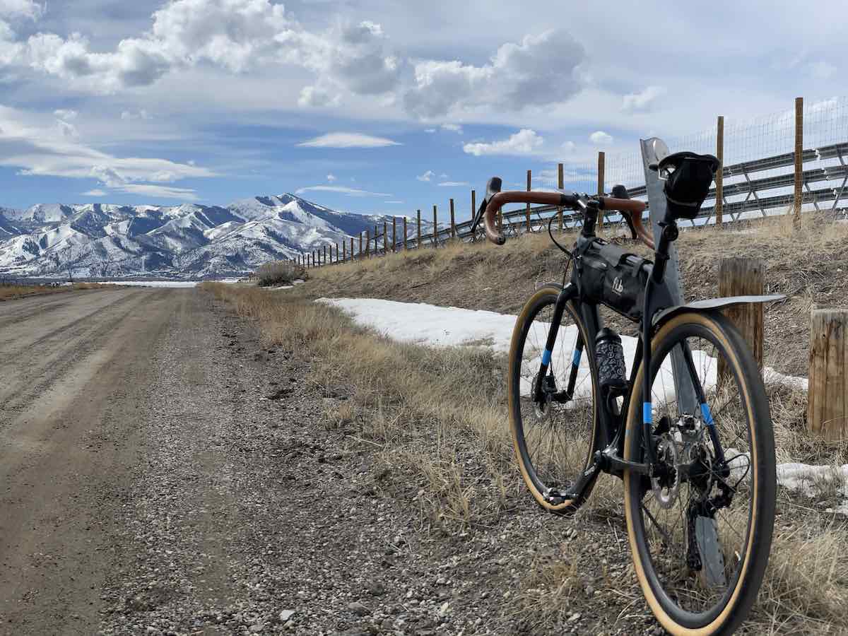bikerumor pic of the day a gravel bike is in the right of the photo facing towards snow capped mountains in the distance. there is a wood fence bordering the gravel road and fluffy white clouds in the sky.