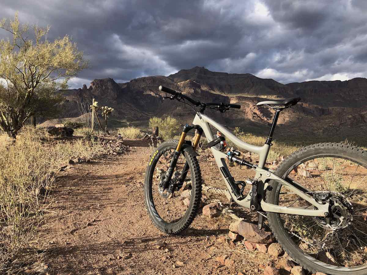 bikerumor pic of the day mountain bike at the Superstition mountain foothills near Phoenix arizona a full suspension mountain bike is perched on the edge of a rocky trail with scrub brush and mountain in the distance sun is peeking through the heavy clouds in the sky