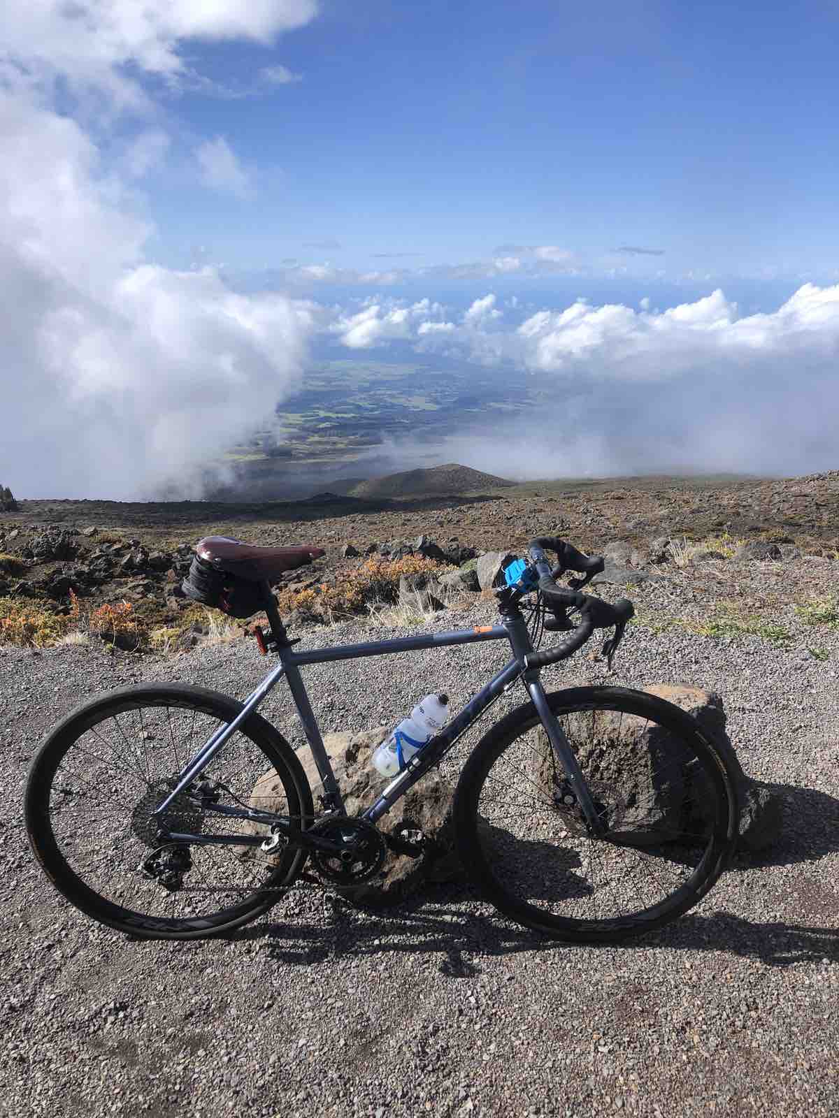 a bicycle is leaning against a rock a the top of haleakala in hawaii. The view looks out over the island through the clouds.