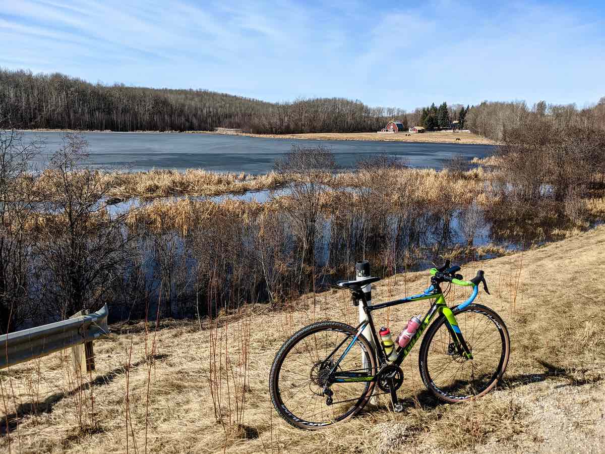 bikerumor pic of the day a gravel bike is positioned in a golden grass area near a lake - across the lake there is a barn. the day is bright and there are whips clouds in the sky.