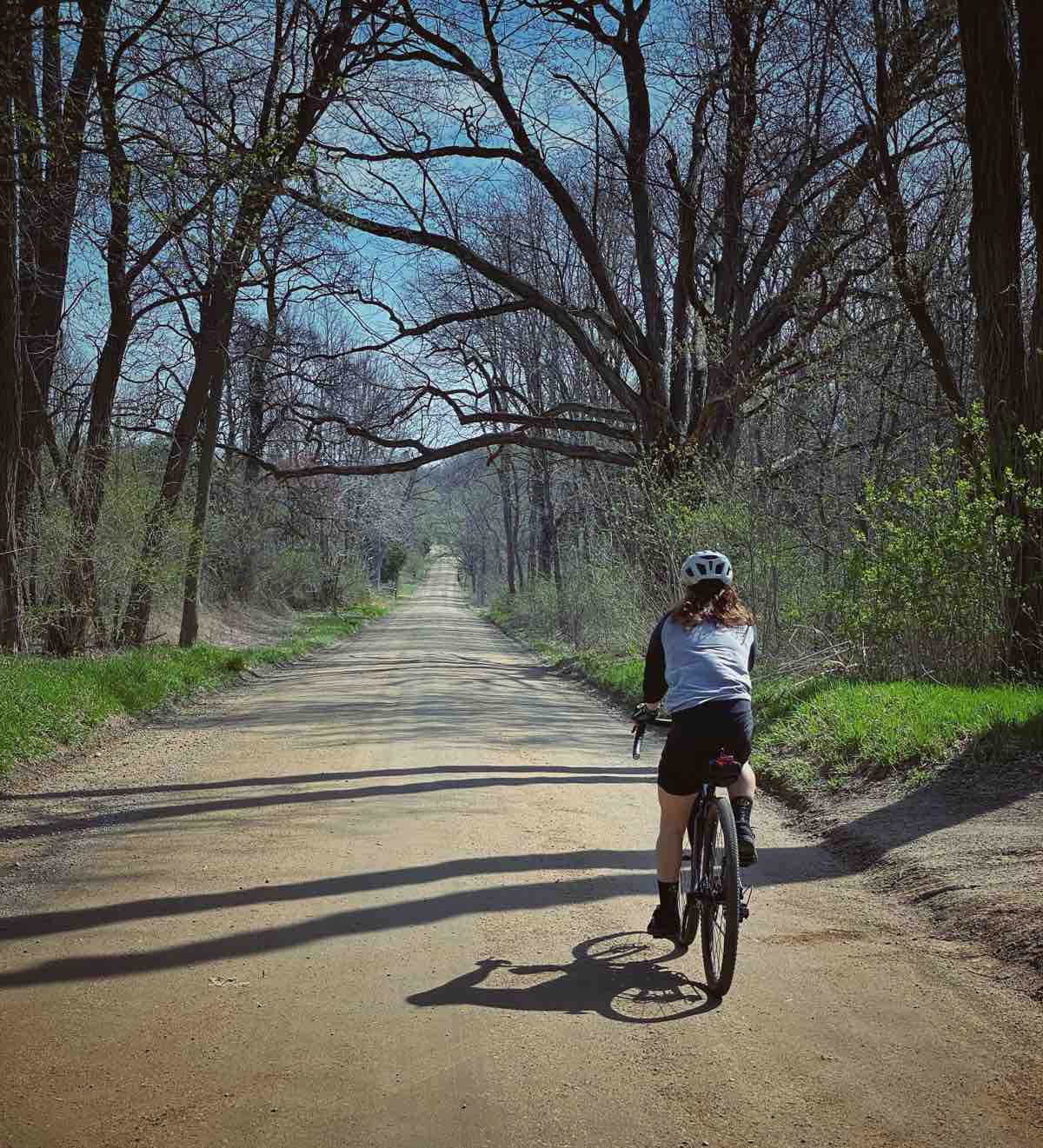 a bicyclist rides on a packed dirt road facing away from the camera. There are trees on either side of the road that are just beginning to leaf out and grass is very green on the edges.