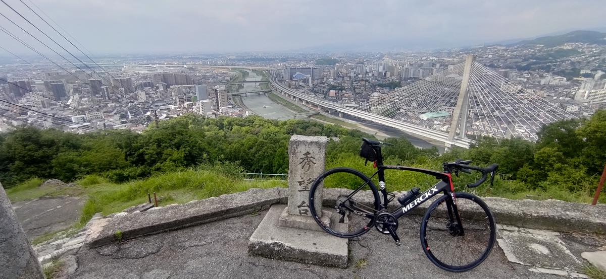bikerumor pic of the day Ikeda city, Osaka, a bicycle is posed near a stone marker at the top of a mountain overlooking a city with hazy mountains in the distance.