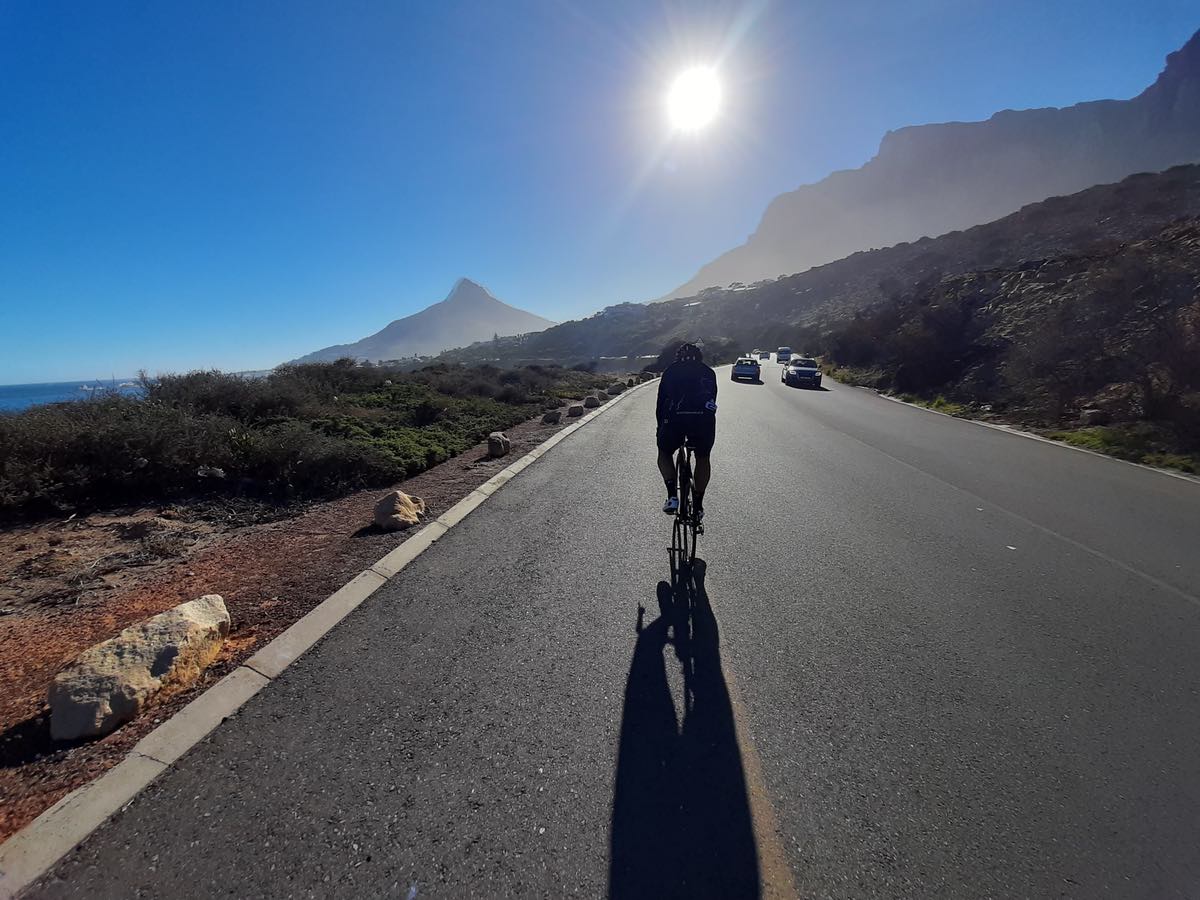 bikerumor pic of the day a cyclist is riding on a paved road away from the camera towards the sun that is low in the blue sky and some mountain peaks in the distance.