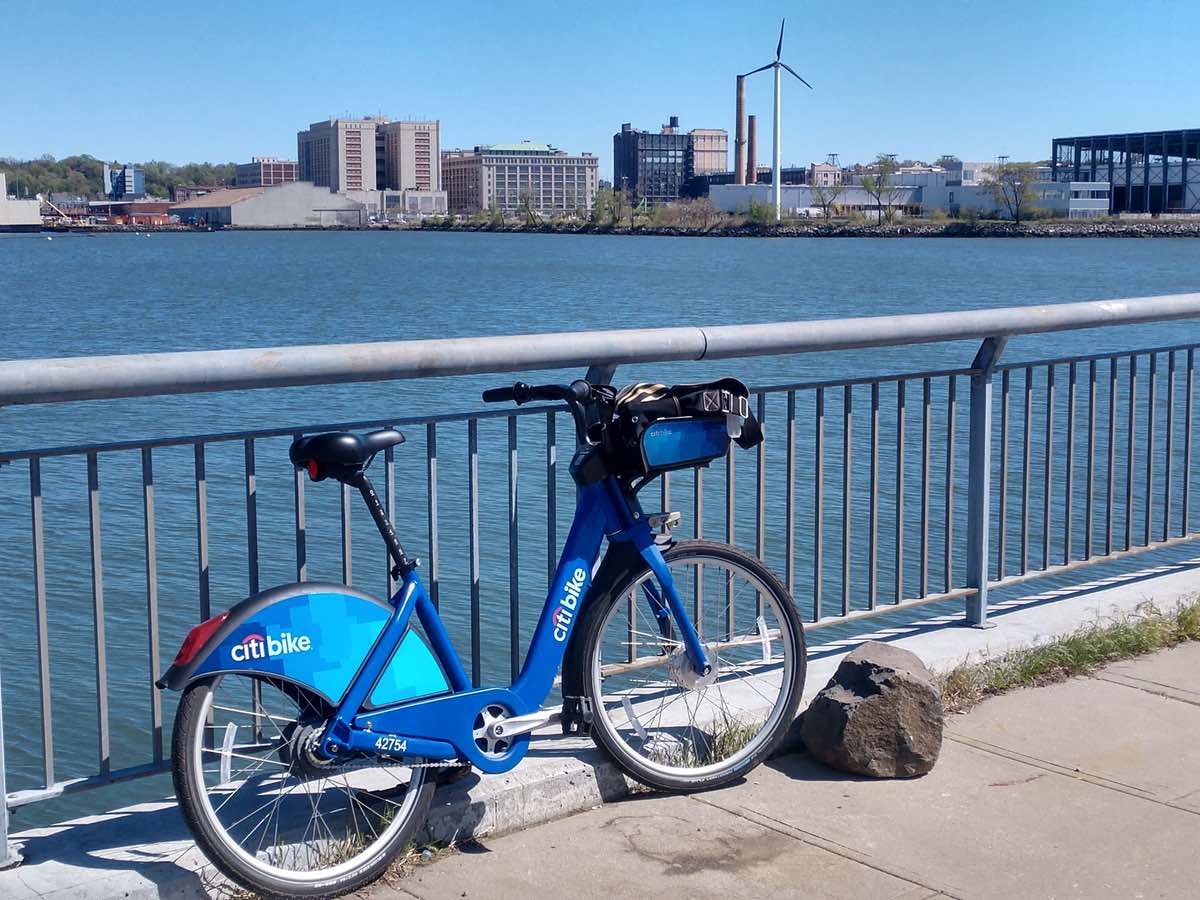 bikerumor pic of the day a citibike leans against a railing along a path overlooking city buildings and a wind turbine in brooklyn new york.