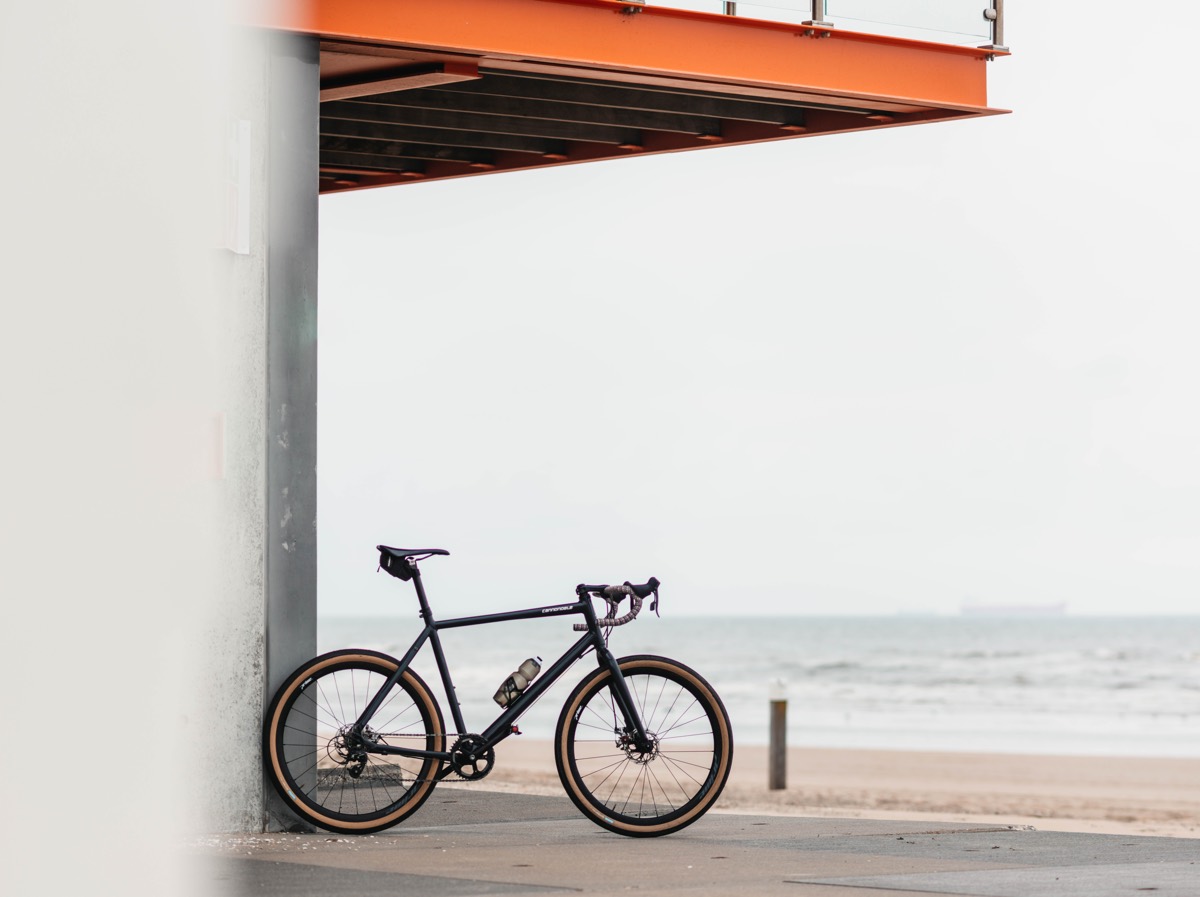 bikerumor pic of the day a bicycle leans against a wall under an orange steel platform at the sea. the sea is grey and the sky looks very white and cloudy.