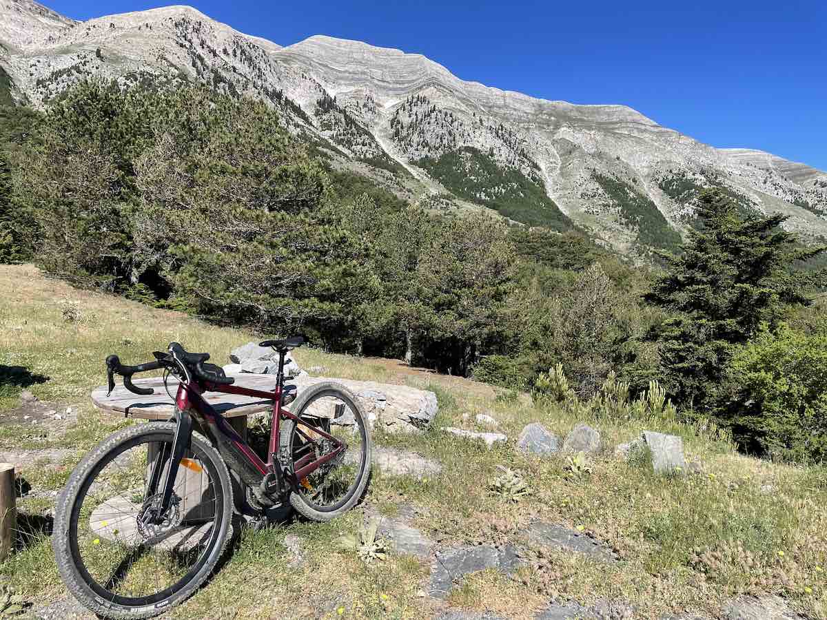 bikerumor pic of the day a gravel bike is perched on the side of Mount Taygetos among the grass and rock outcropping with pine trees and the mountain in the background, the sky is clear blue and it is very sunny
