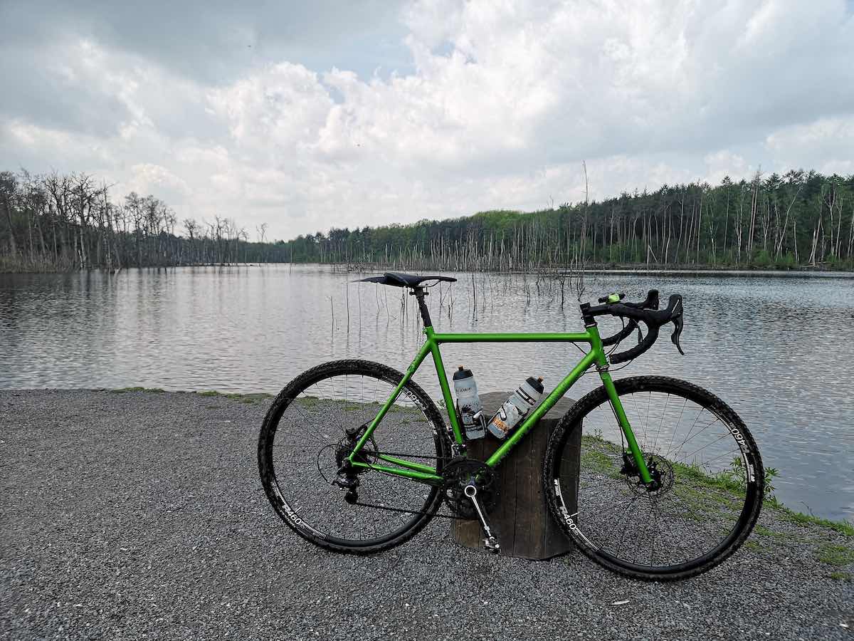 bikerumor pic of the day a green grave bike is on a gravel beach on the edge of a body of water with cloudy sky reflecting in it