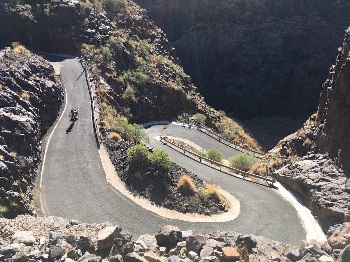 bikerumor pic of the day cyclist is riding a bike with a child trailer carrier down a curvy and windy road in gran canaria spain it is very sunny and the sides of the road are rocky.