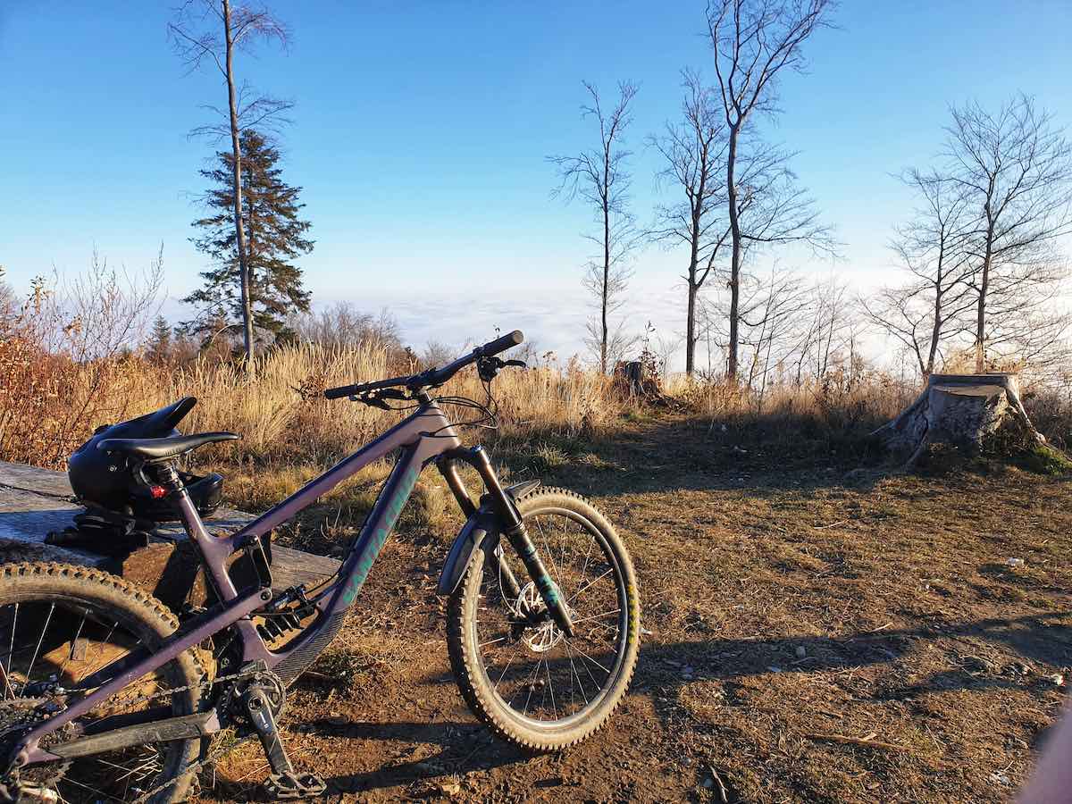 bikerumor pic of the day a mountain bike leans against some logs at the top of sljeme mountain the sun is low and golden on the packed dirt and brush the view is of the clouds over the city of zagreb croatia.