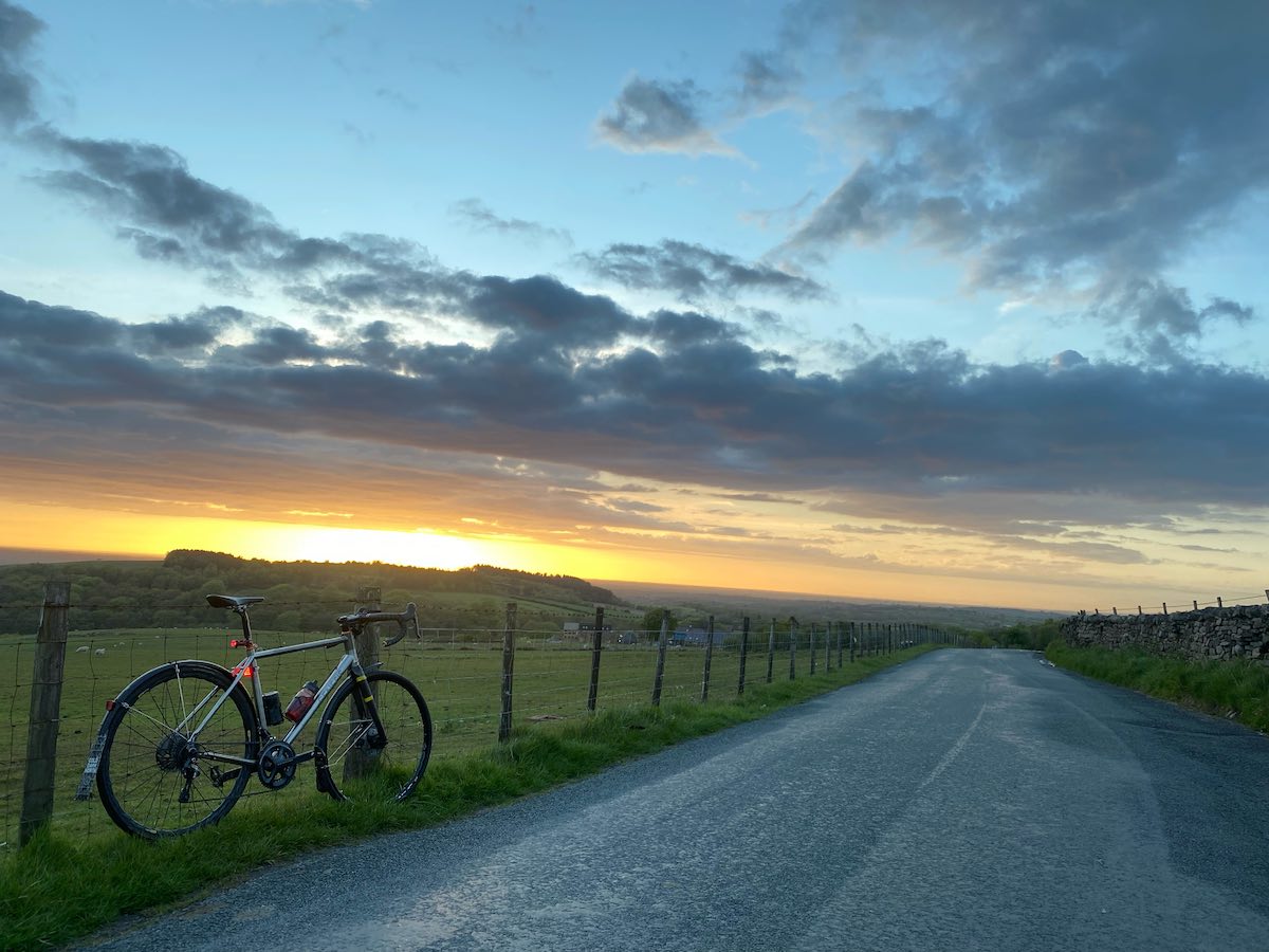 bikerumor pic of the day a bicycle leans against a wire fence along a gravel road next to large green fields, the sun is setting and the clouds and sky are very colorful.
