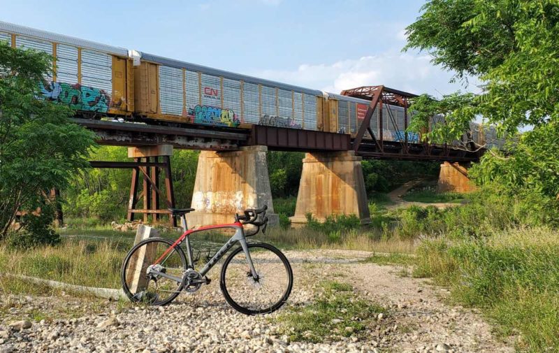 bikerumor pic of the day a bicycle is on a gravel path near a train bridge, there is colorful graffiti on the train cars and scrubby trees on either side of the path.
