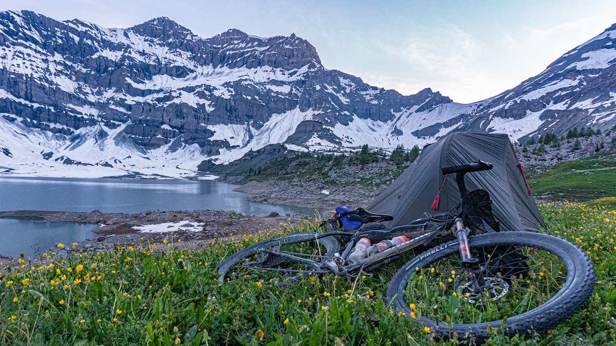 bikerumor pic of the day a bicycle lays in the grassy field next to a tent overlooking a lac with snow speckled mountains in the background.