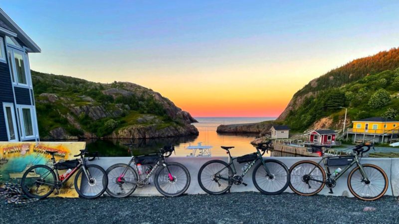 bikerumor pic of the day four bicycles lean against a concrete barrier on a gravel road, there is a small harbor in the background with small colorful buildings by the water, the sky is orange at the horizon and is reflecting on the water, the sky is clear.