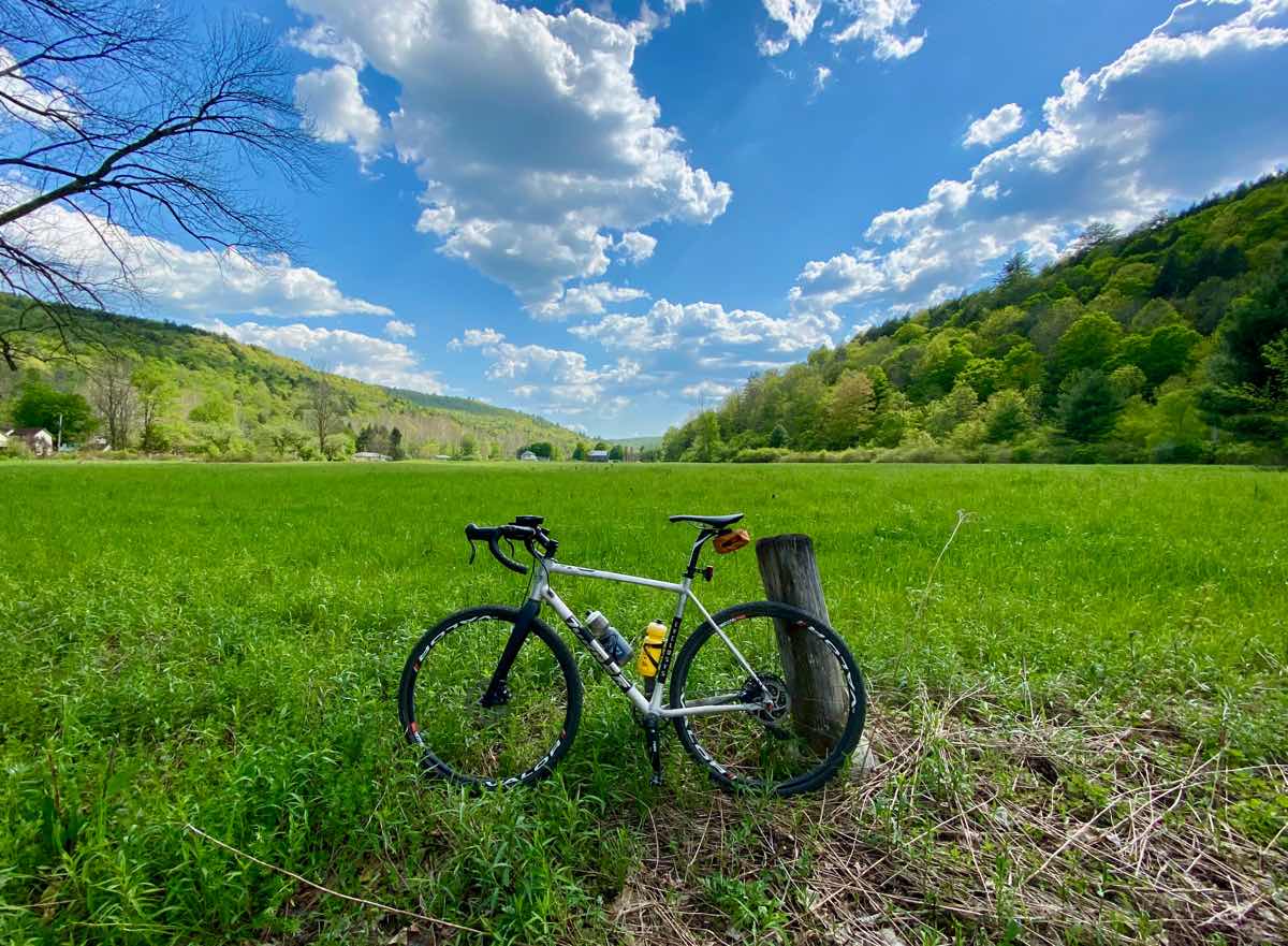 bikerumor pic of the day a bicycle leans against a short wood post in a bright green grassy field surrounded by mountains with bright blue sky filled with fluffy clouds