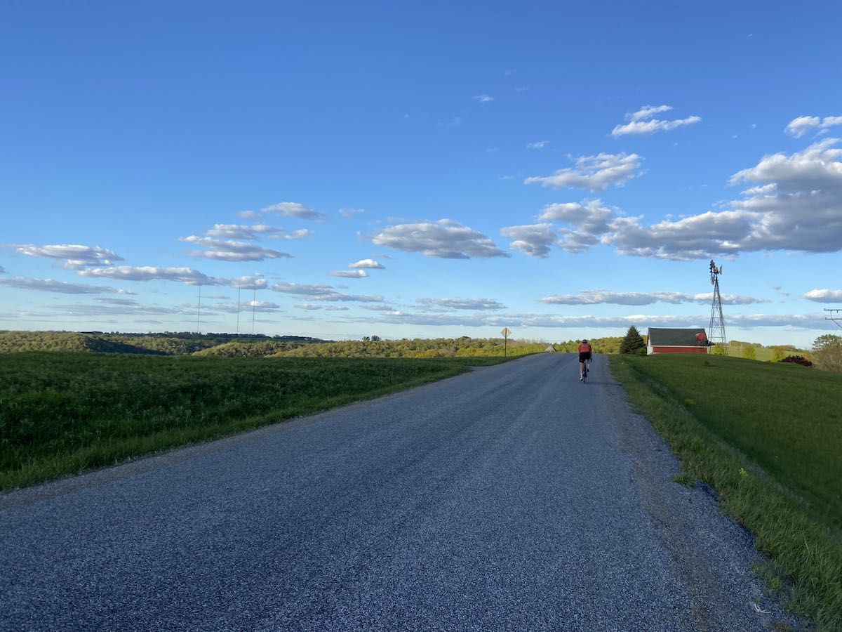 bikerumor pic of the day a cyclist is riding away from the camera on a road that is surrounded by green fields a small red barn and windmill can be seen just past the rider and there are tree-filled hills beyond that. the sky is blue and there are a few clouds in the sky at what looks like evening.