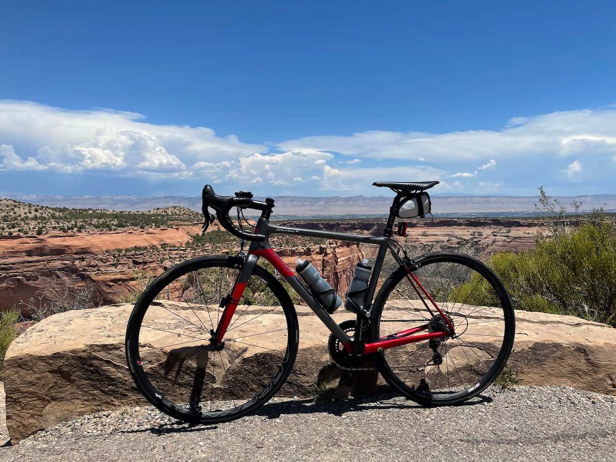 bikerumor pic of the day a bike is atop a mesa overlooking colorado national monument there is red dirt, rocks and low scrub trees the sun is high in the sky and it is bright blue with white clouds in the distance