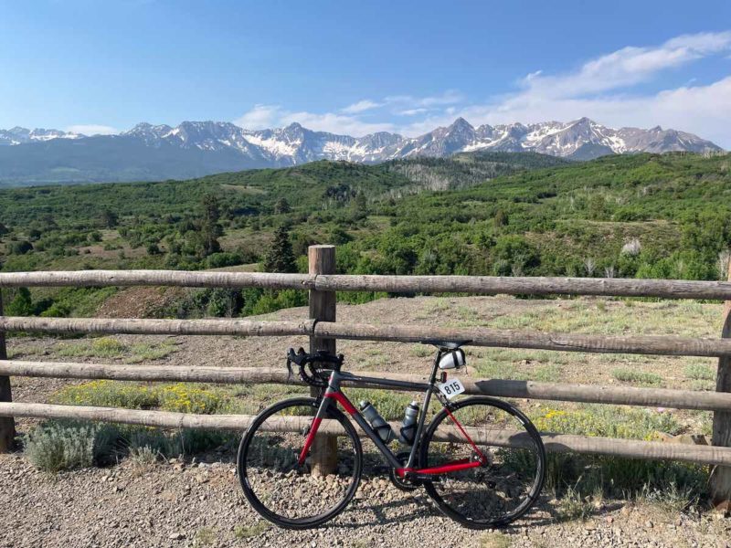 bikerumor pic of the day a bicycle leans against a wooden fence on a gravel road, a grassy field is next to it with tree covered hills and rocky mountains beyond, the sky is blue with a few clouds on the horizon.