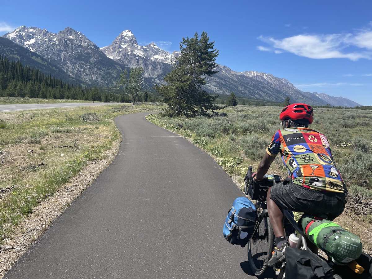 bikerumor pic of the day a cyclist rider a road surrounded by grassland leading towards a large pointed snow speckled mountain in the grand teton national park, wyoming.