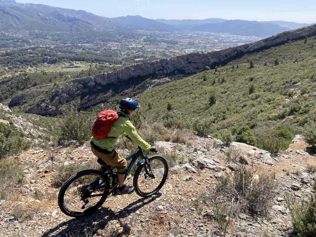 bikerumor pic of the day a mountain biker is on the edge of a trail overlooking rocky outcroppings on the mountain and a valley below, the sun is high and the sky is clear.