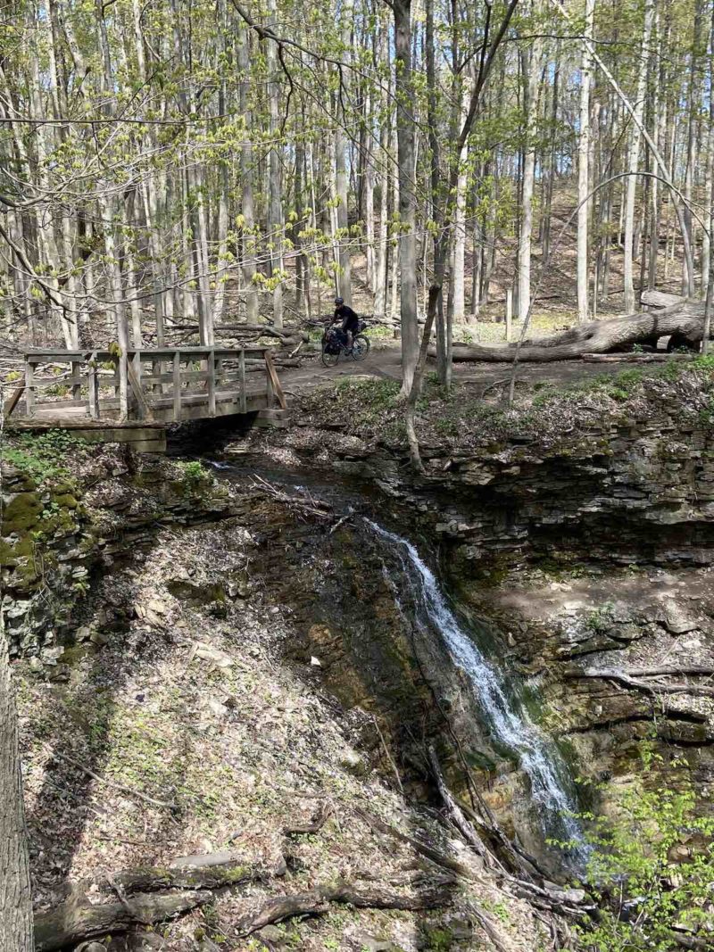bikerumor pic of the day a cyclist is on a trail that edges the woods on one side and a rocky edge with a waterfall on the other a small bridge is before them, sunlight is coming through the trees and leaves are just beginning to open on the trees.