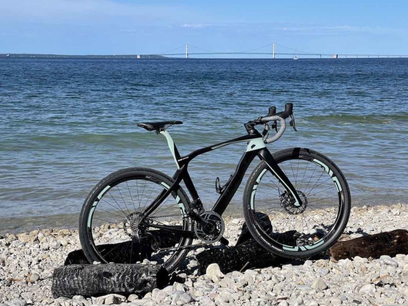 bikerumor pic of the day a bicycle lis on a beach with a long suspension bridge in the distance, the sky is clear and the sun is bright.