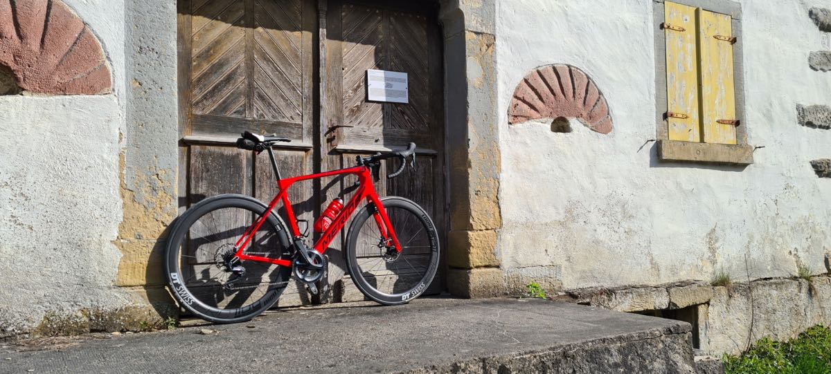 bikerumor pic of the day a red road bike leans agains the large wooden doors of an old mill. the building is of white stucco with stone accents and appears to be very old.