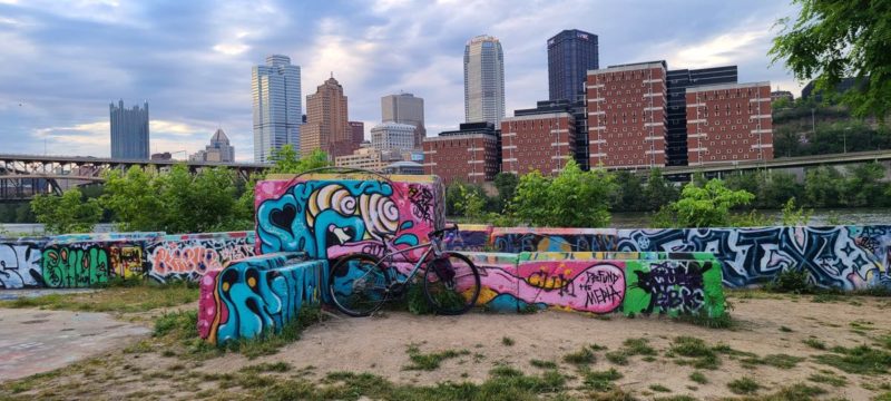 bikerumor pic of the day a bicycle is in front of some very colorful graffiti with the skyline of pittsburgh pa in the background the sky is cloudy with bits of blue and light from the sun peeking through.