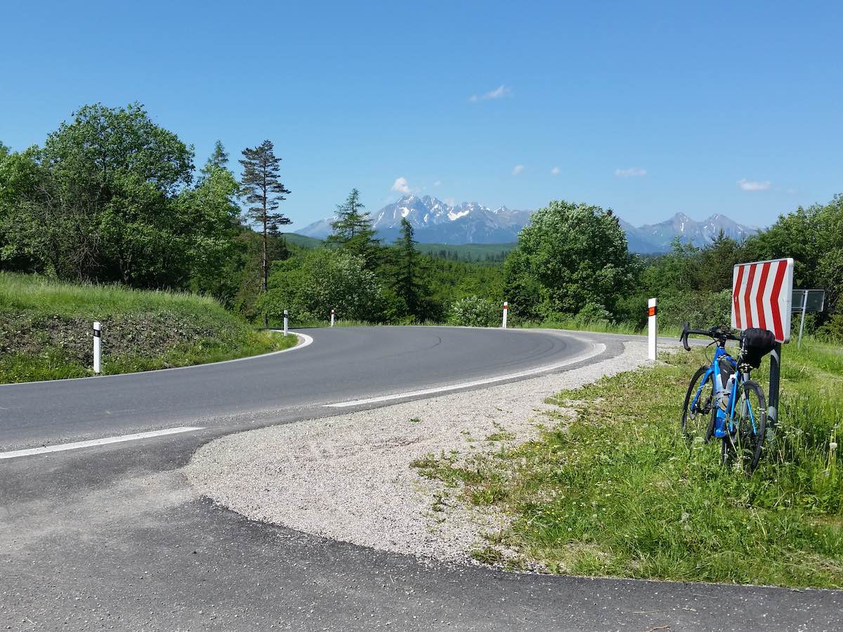bikerumor pic of the day a road bike leans against a red and white road sign with a high mountain pass in the distance. The sky is clear and blue and there are tall trees along the road.