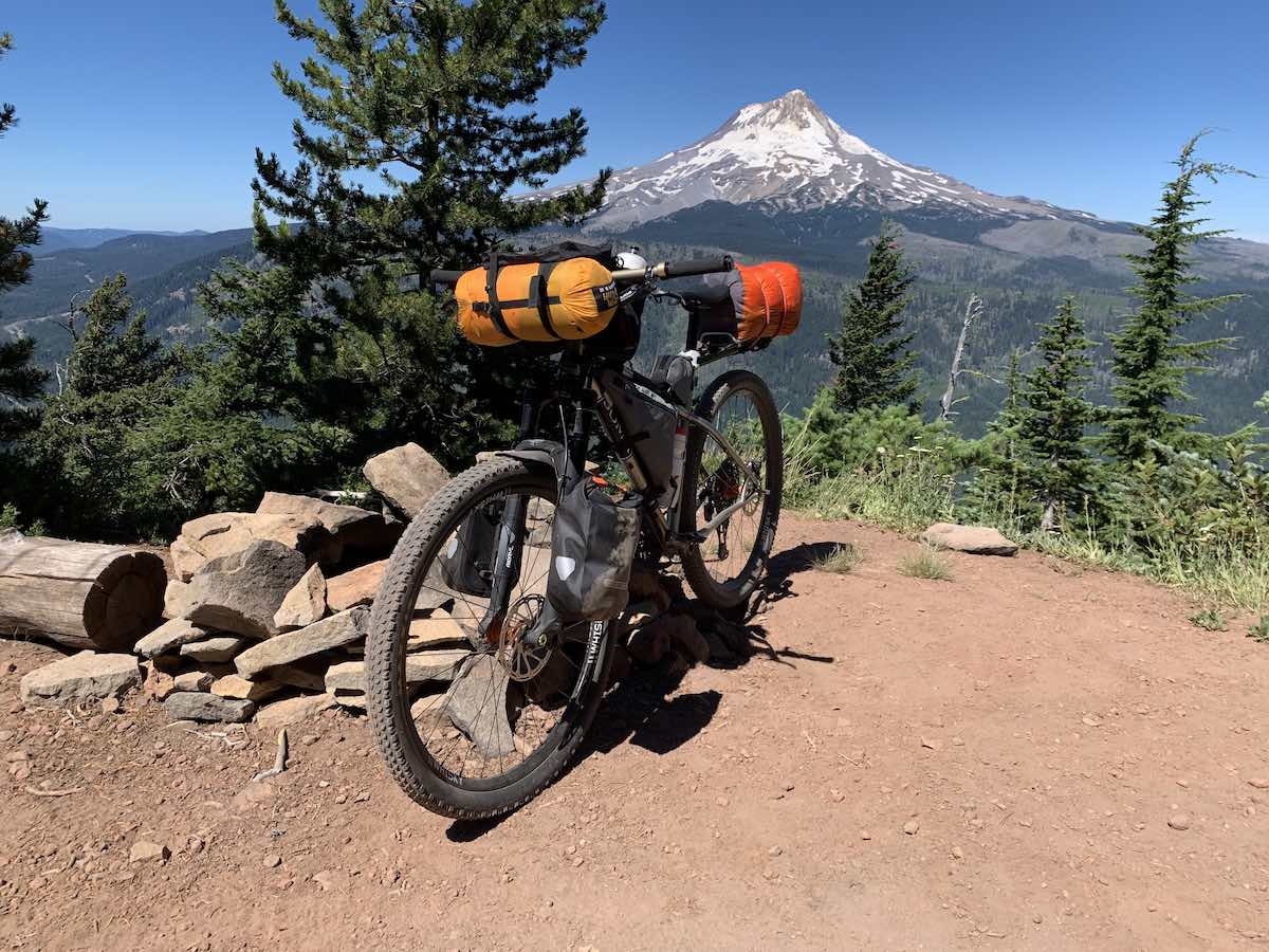 bikerumor pic of the day a bicycle with camping gear packed on it is on a clearing with pine trees surrounding it and a view of a snow capped mountain peak in the distance.