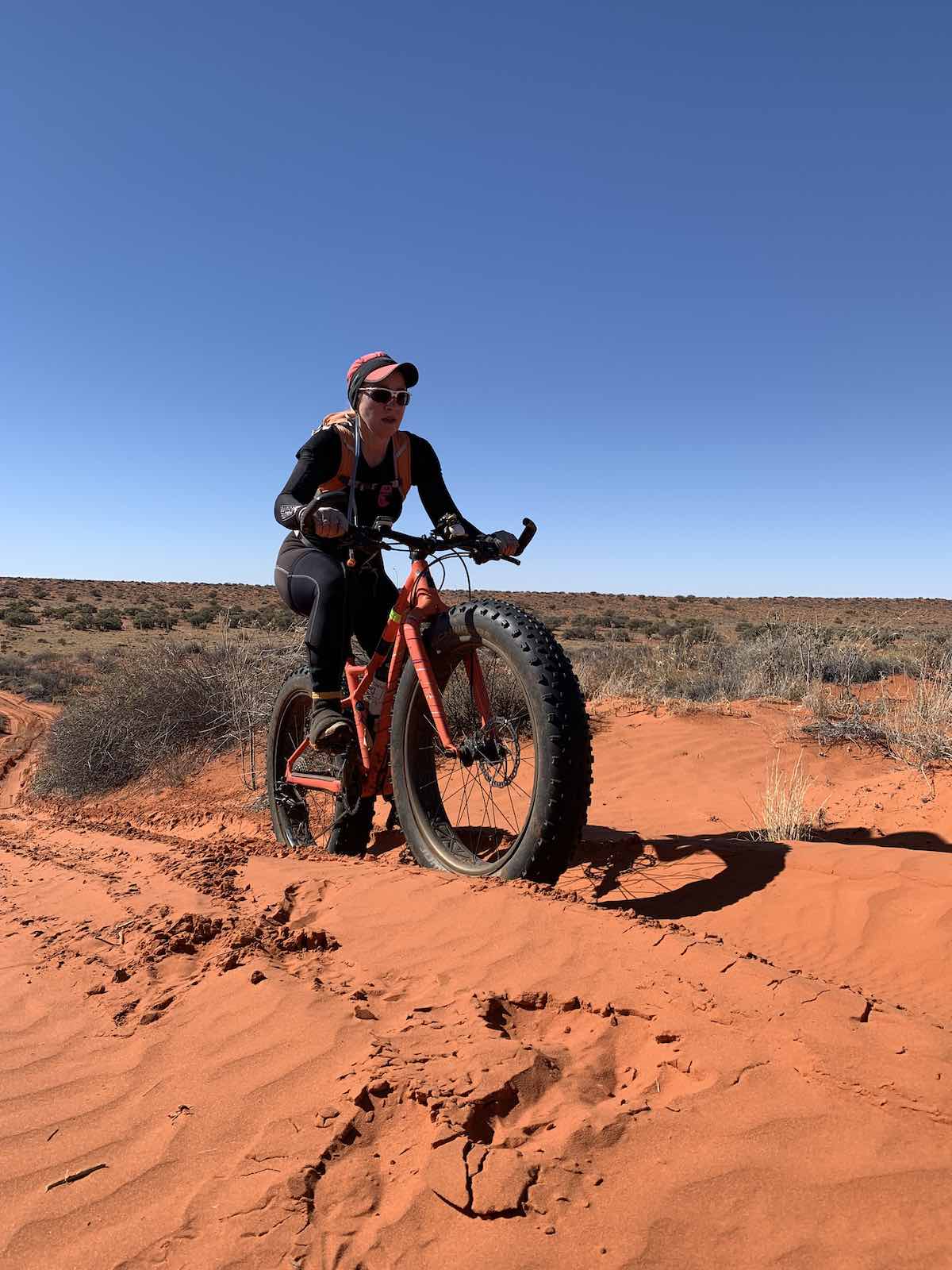 bikerumor pic of the day a cyclist rides a fat tire bicycle on red desert sand, the sky is clear and dark blue, there is brush growing in the flat distance.