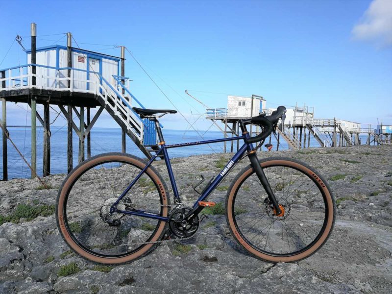 bikerumor pic o the day a bicycle is on a rocky beach in St Palais sur Mer there are freestanding structures on stilts at intervals along the shore the ocean is calm and there are no clouds in the sky.