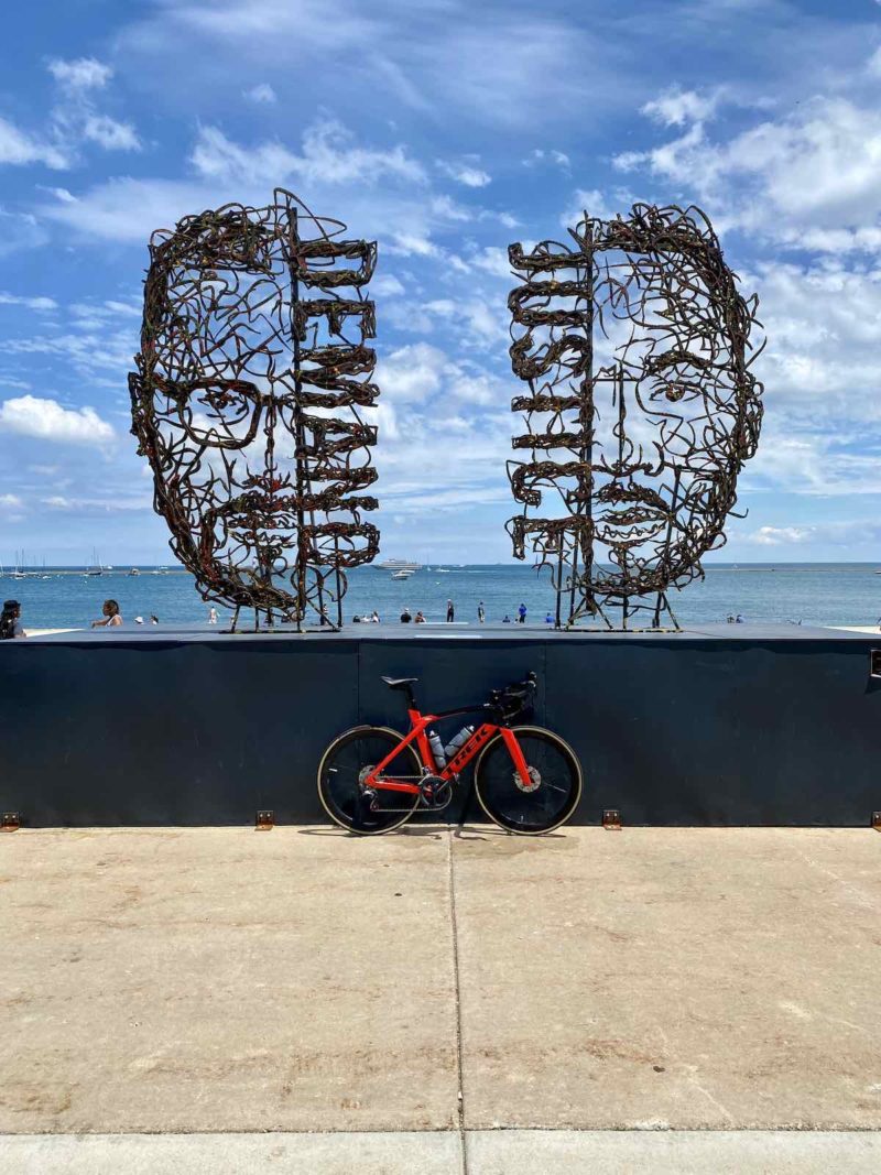 bikerumor pic of the day a red bicycle is leaning against a wall the borders a waterfront, there are two metal sculptures on the wall one is the face of malcom x with the word demand and the other is the face of martin luther king jr with the word justice on one side. the sky is blue with white fluffy clouds interspersed and there are people out by the water.